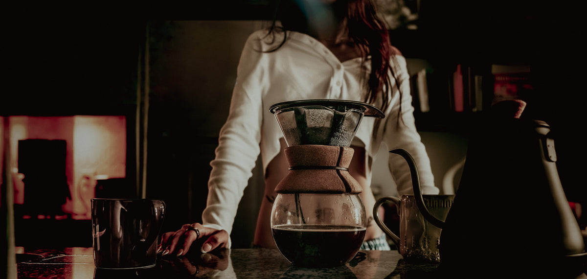 Person making coffee using a pour-over setup in a dimly lit room.